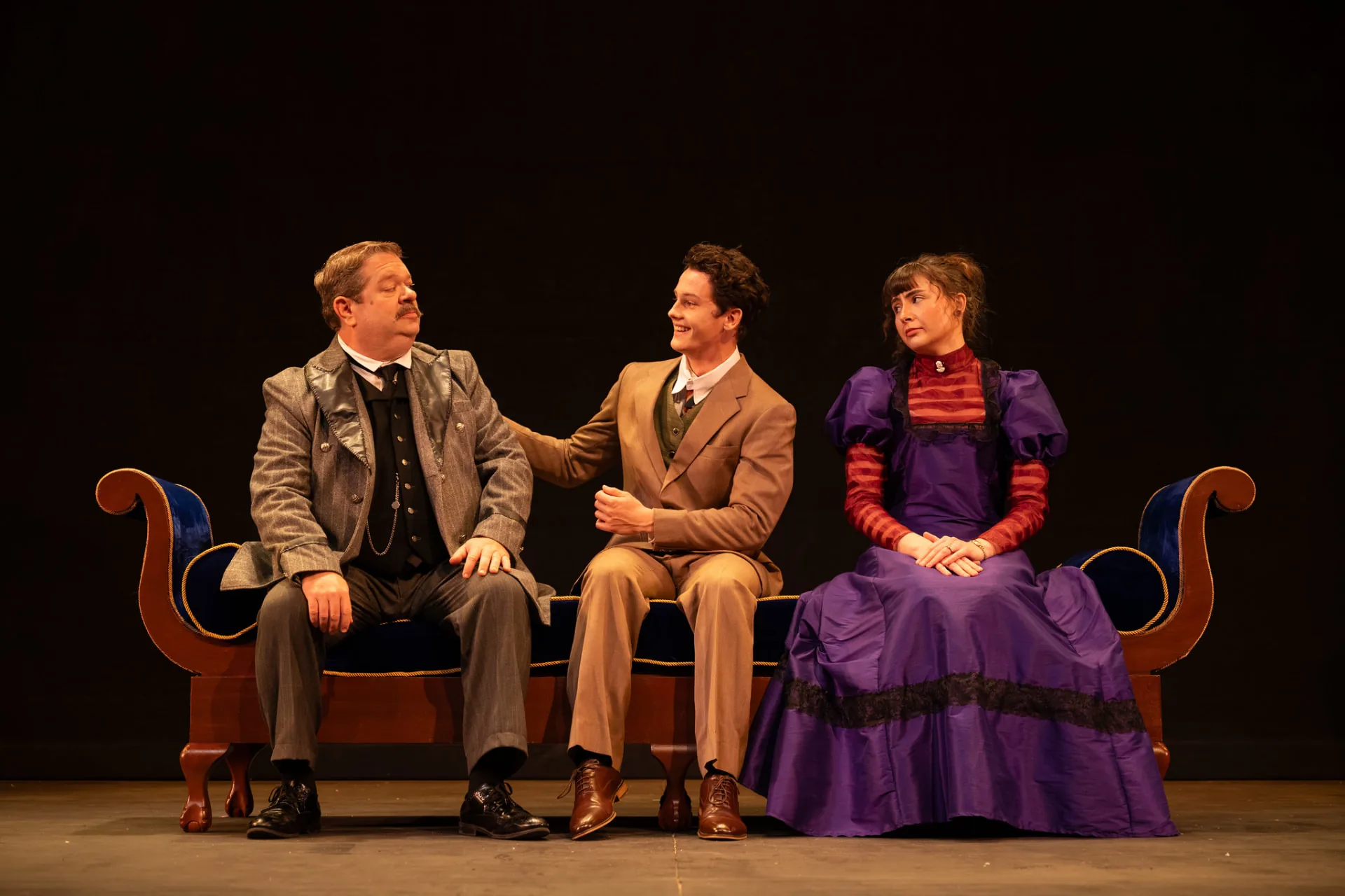 Undershaft seated on a blue velvet chaise as a young man lays a hand on his shoulder and a young woman in purple looks on