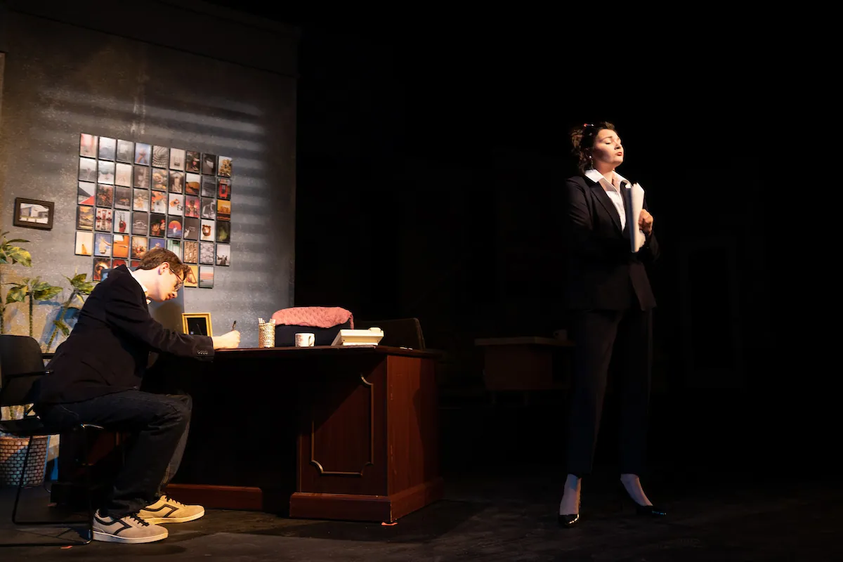 A woman in a black suit stands as a man works at a desk beneath a wall of photographs
