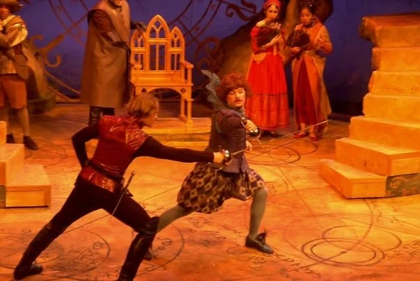 Jim Harris as Astolfo, in a curly red wig and quilted doublet, crossing rapiers with a man in a red jerkin