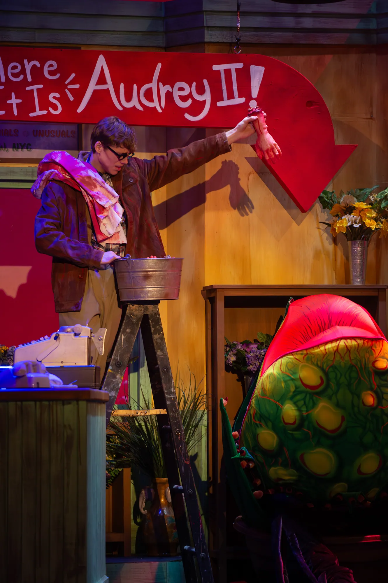 A performer beside the plant beneath a banner reading Here It Is, Audrey II