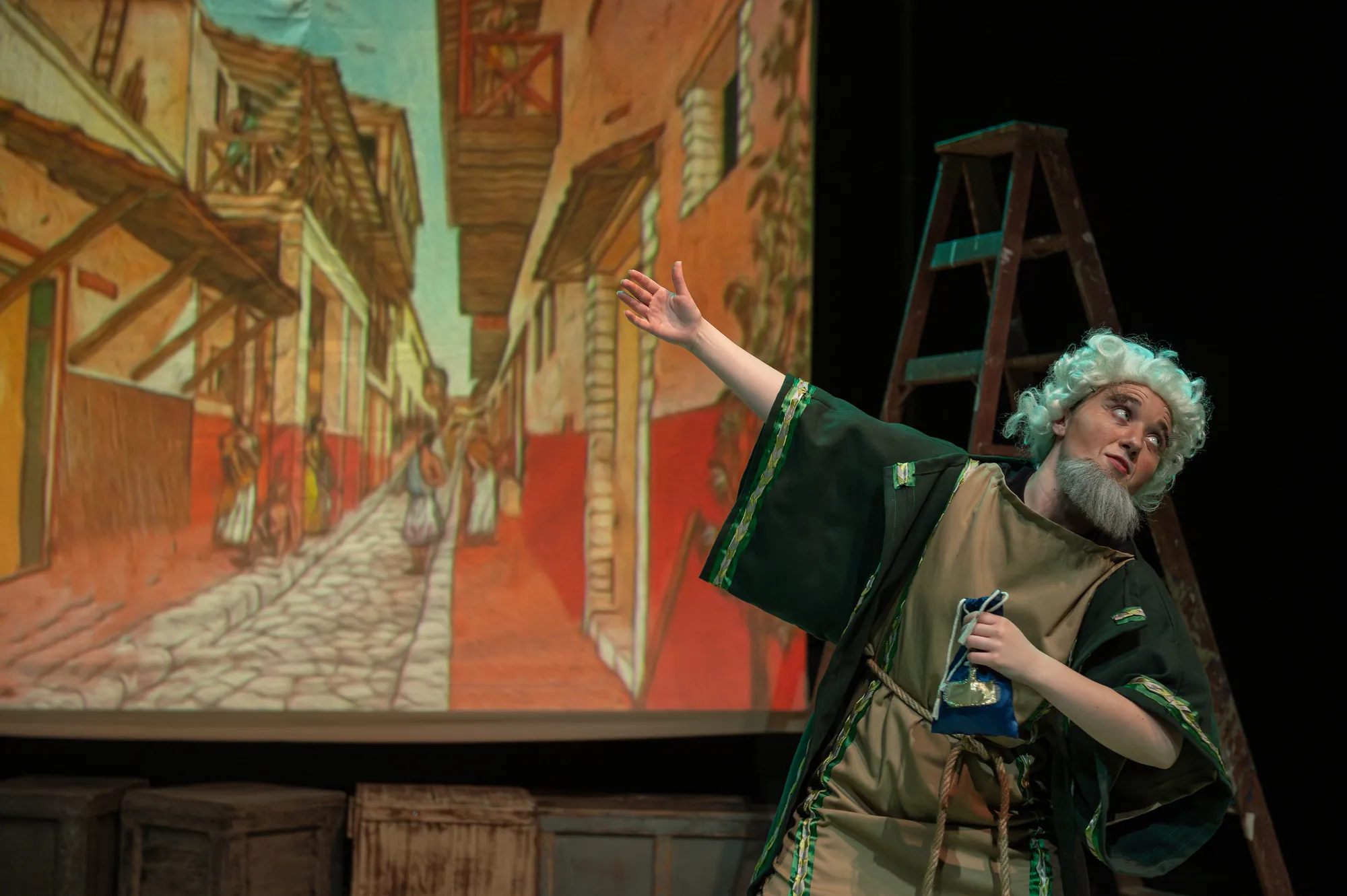A bearded old man in a robe gestures before a painted street scene