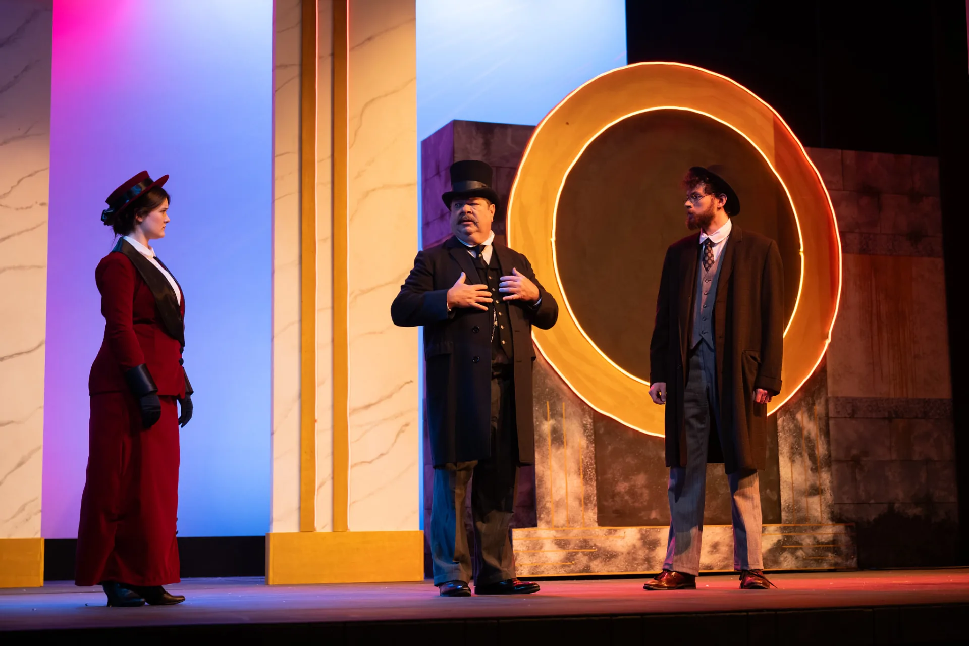 Undershaft, hand on his chest, between a woman in a red suit and a bearded young man in a bowler hat