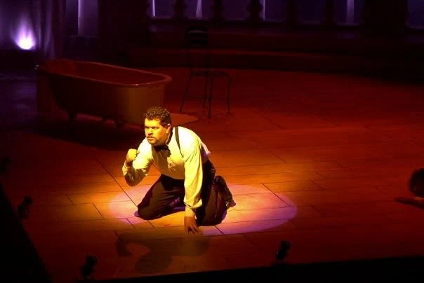 A young Jim Harris as Constantine, kneeling in a pool of amber light with a clenched fist, a clawfoot bathtub behind him