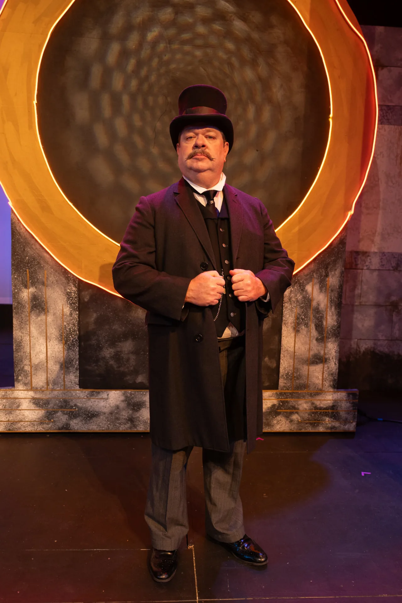 Jim Harris as Undershaft in top hat and frock coat, gripping his lapels before a glowing gold ring