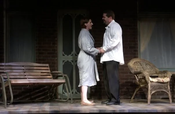 Hal and a woman in a bathrobe hold hands and smile at each other on the porch