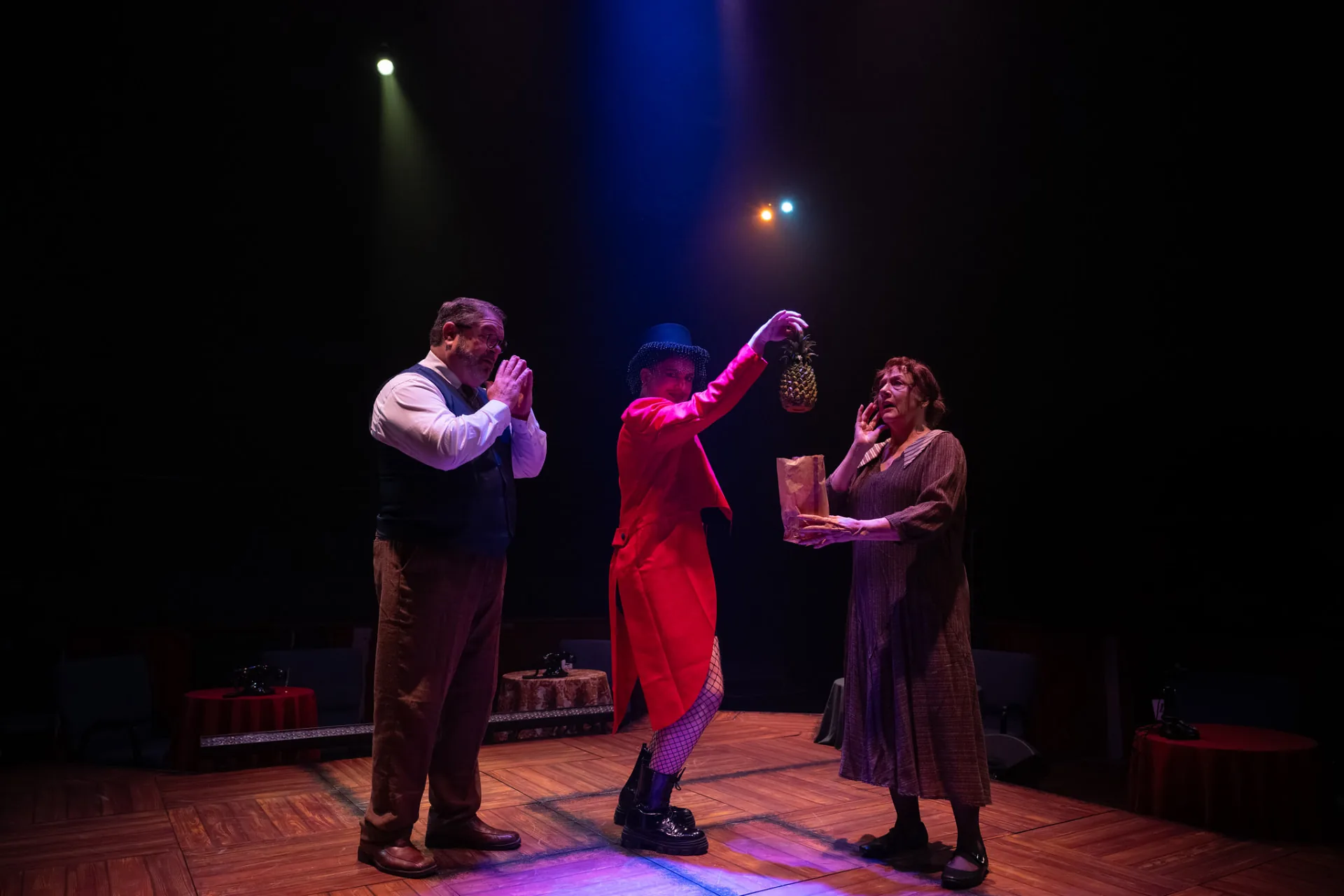 The Emcee in a red tailcoat dangles a pineapple between Herr Schultz, hands pressed together, and a startled woman