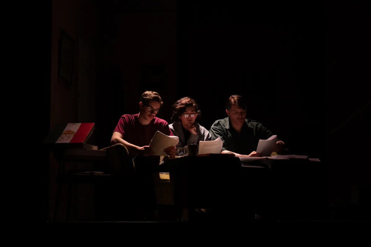 Three actors read together at a table in a pool of light
