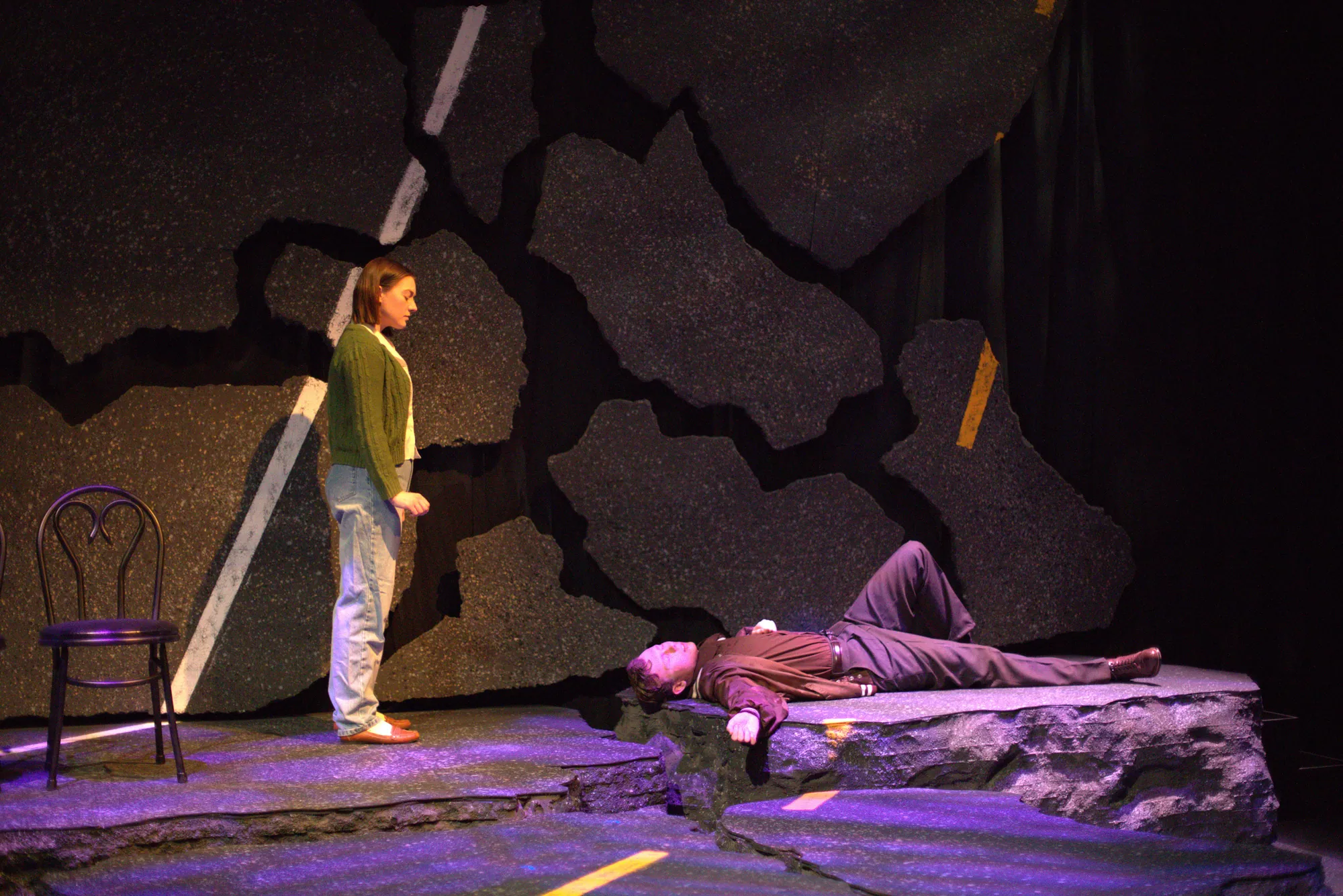 A woman on the highway set as a man lies beside a chair in purple light