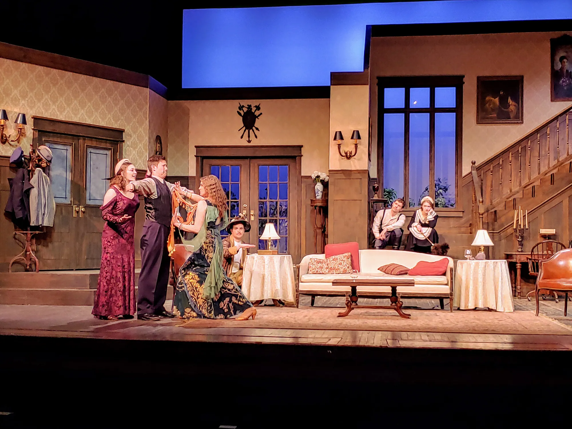 The company crowds around a woman in the drawing room