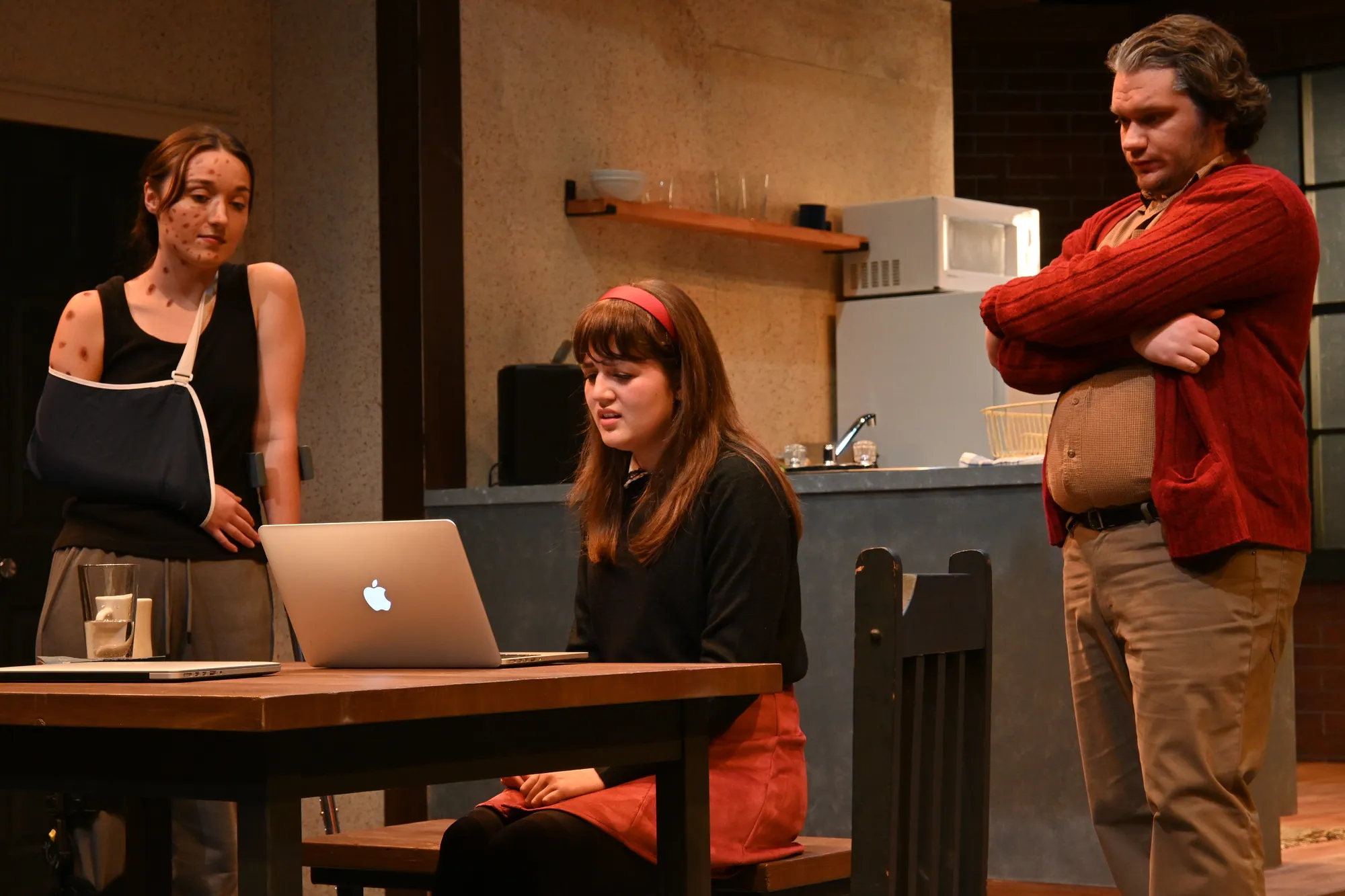 A woman with her arm in a sling sits at a laptop while a man looks on