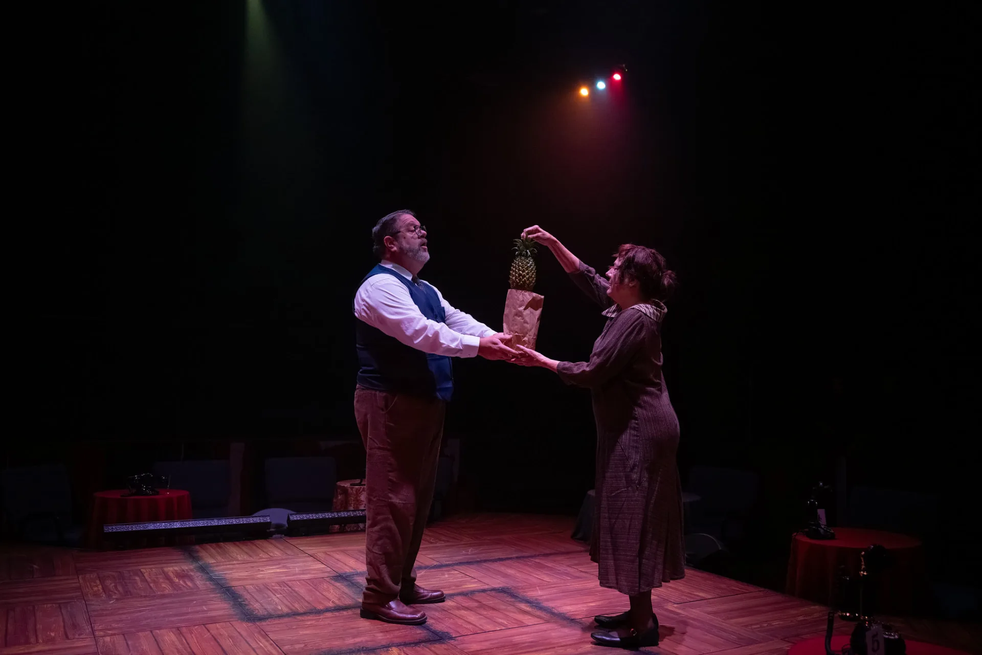 Herr Schultz and a woman hold a pineapple in a paper bag between them on a wooden dance floor